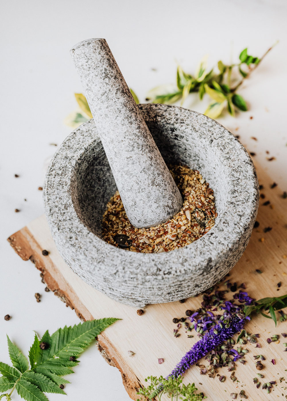 A stone mortar and pestle filled with seeds and surrounded by different edible plants and flowers