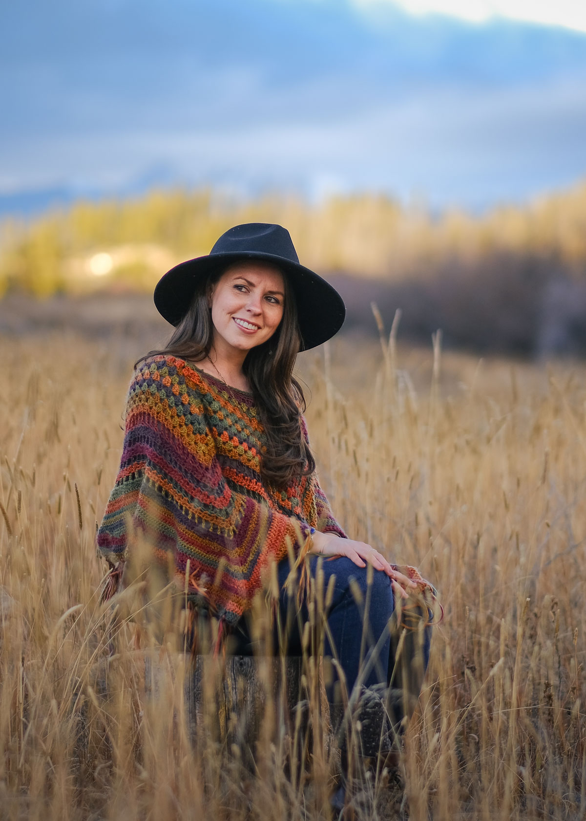 Lindsay Knecht, NTP and holistic nutritionist looks off to the side as she sits in a wheat field wearing a knit poncho and outdoorsy fedora