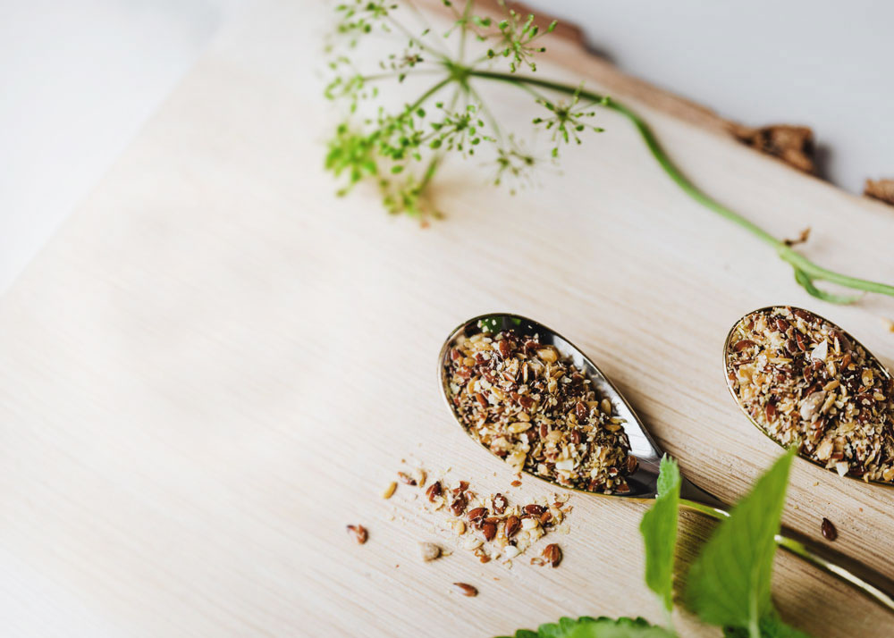 Two spoons overflowing with seeds and several full herbs are set upon a wooden cutting board